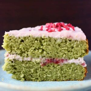 A slice of matcha sponge cake with strawberry frosting on a blue plate against a dark brown background