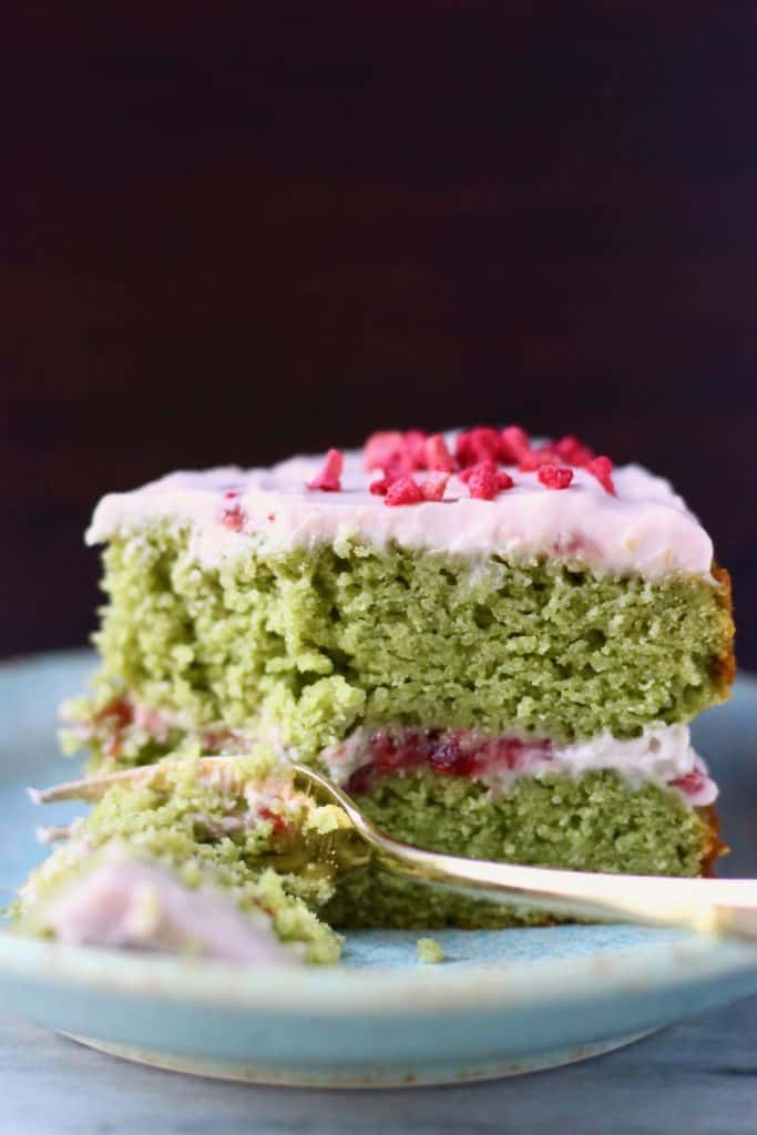 A slice of matcha sponge cake with strawberry frosting on a blue plate against a dark brown background with a gold fork taking a mouthful