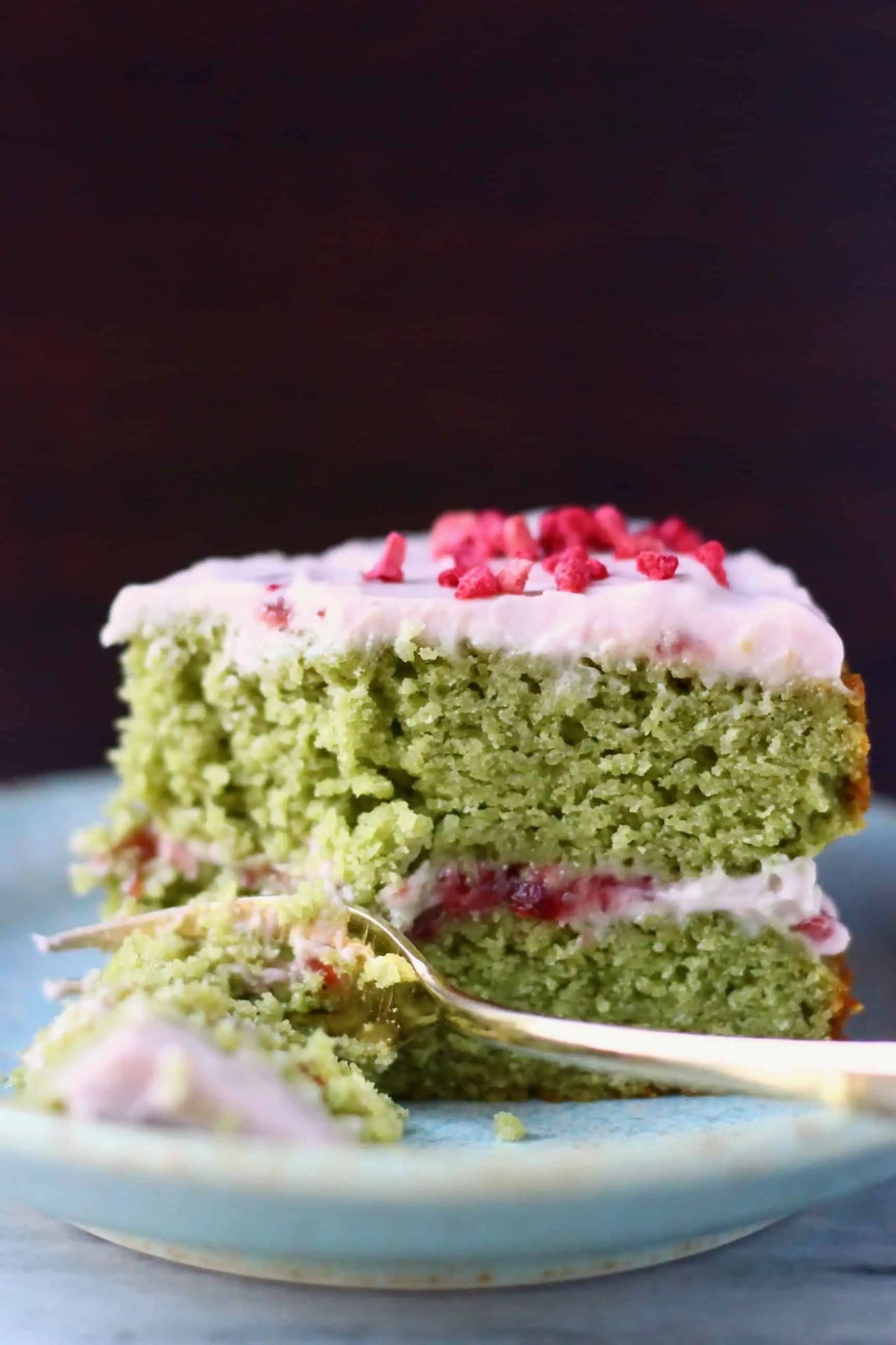 A slice of gluten-free vegan matcha strawberry cake with strawberry frosting on a plate with a fork taking off a mouthful 