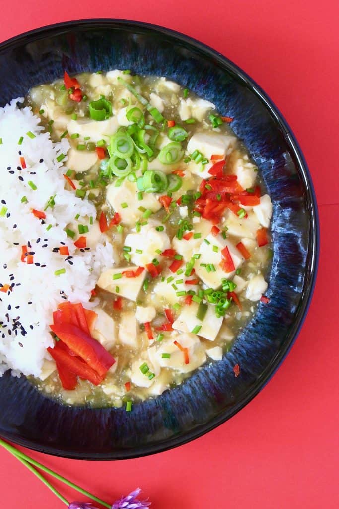 Scrambled tofu in a brown sauce with a pile of rice sprinkled with black sesame seeds in a black bowl against a red background