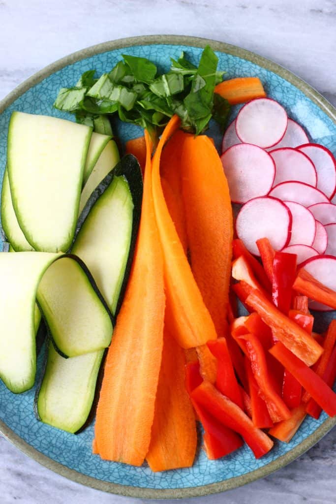 Carrot ribbons, courgette ribbons, red pepper matchsticks, sliced radishes and chopped basil on a blue plate against a marble background