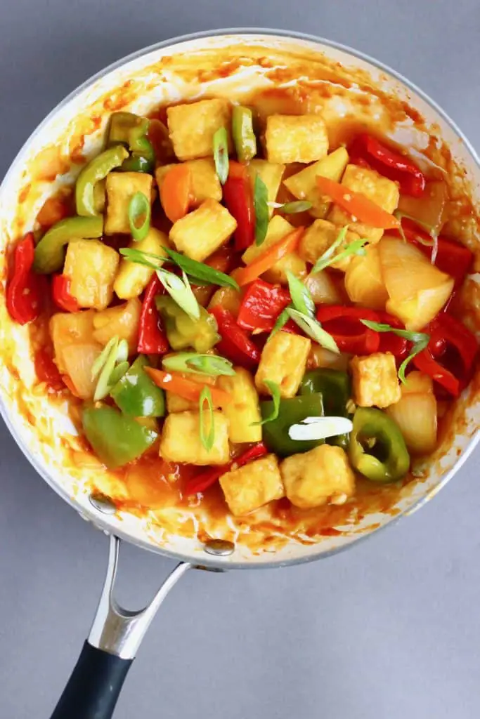 Cubes of tofu with red pepper, green pepper and sliced spring onions in a red sauce in a white frying pan against a grey background