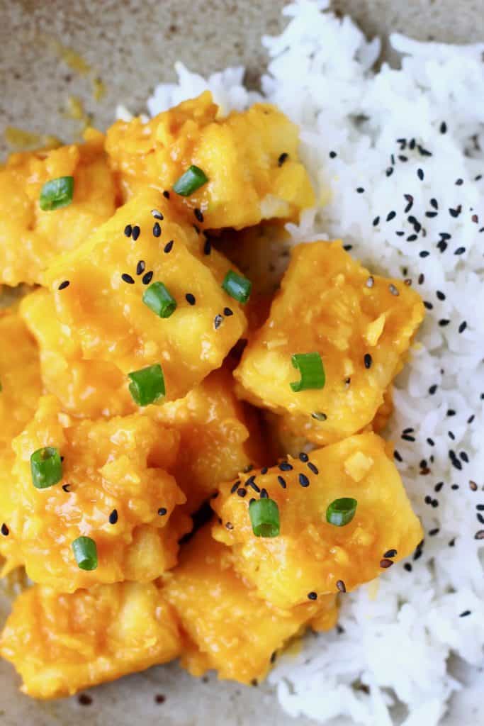 Cubes of tofu covered in orange sauce topped with black sesame seeds and sliced chives with white rice on a light brown plate