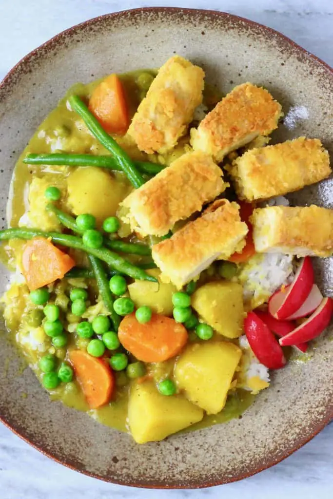 Potatoes, green peas, carrots and green beans in a yellow curry sauce with a pile or rice topped with breaded sticks of tofu on a light brown plate