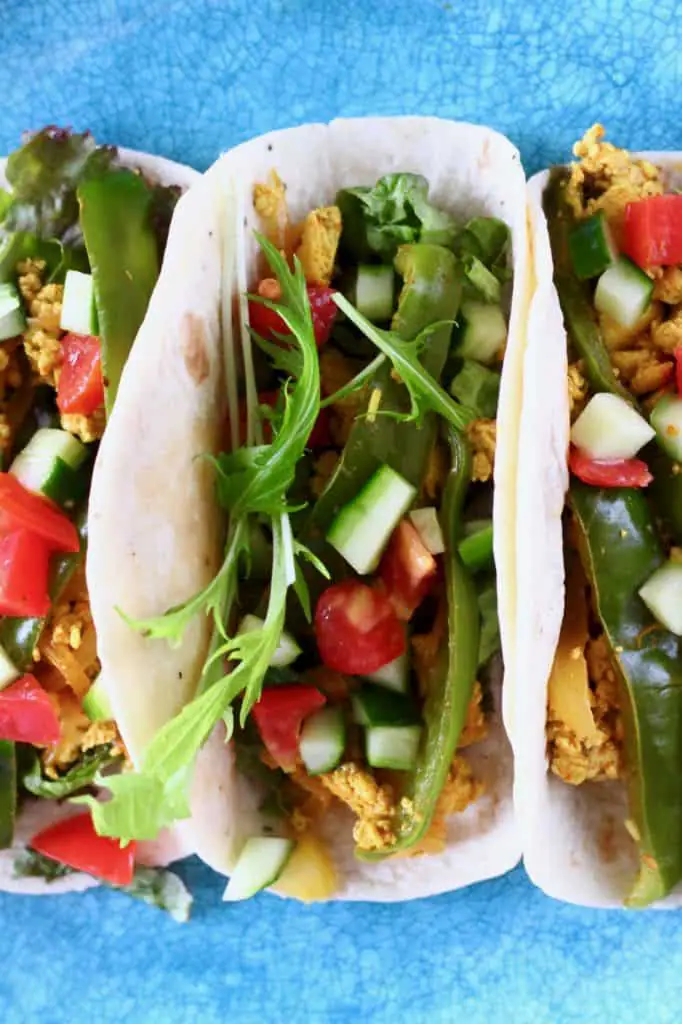 Three tacos filled with scrambled tofu, green peppers and chopped cucumber and tomatoes on a blue plate