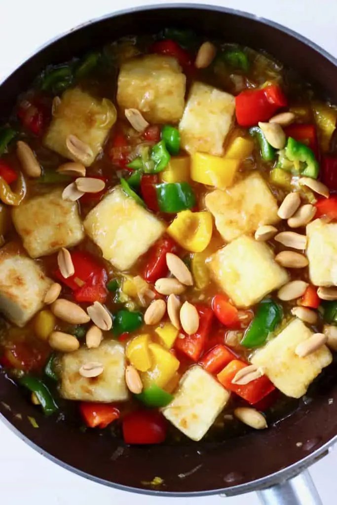 Cubes of fried tofu, red peppers, green peppers and roasted peanuts in a brown sauce in a dark brown frying pan