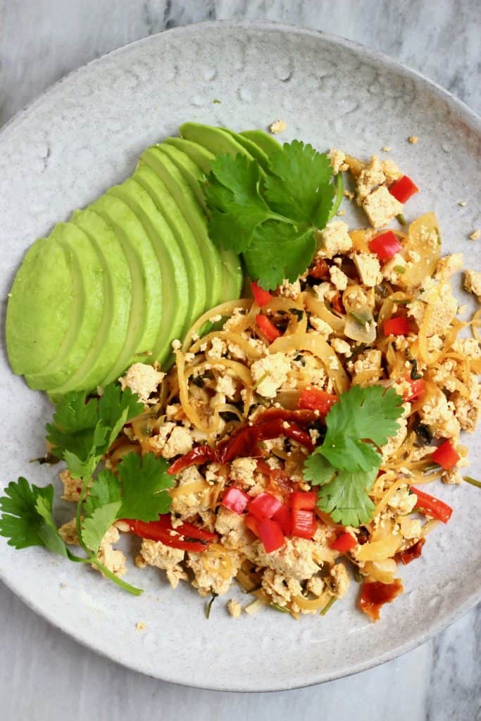 Scrambled tofu with red peppers and a sliced avocado on a grey plate against a marble background