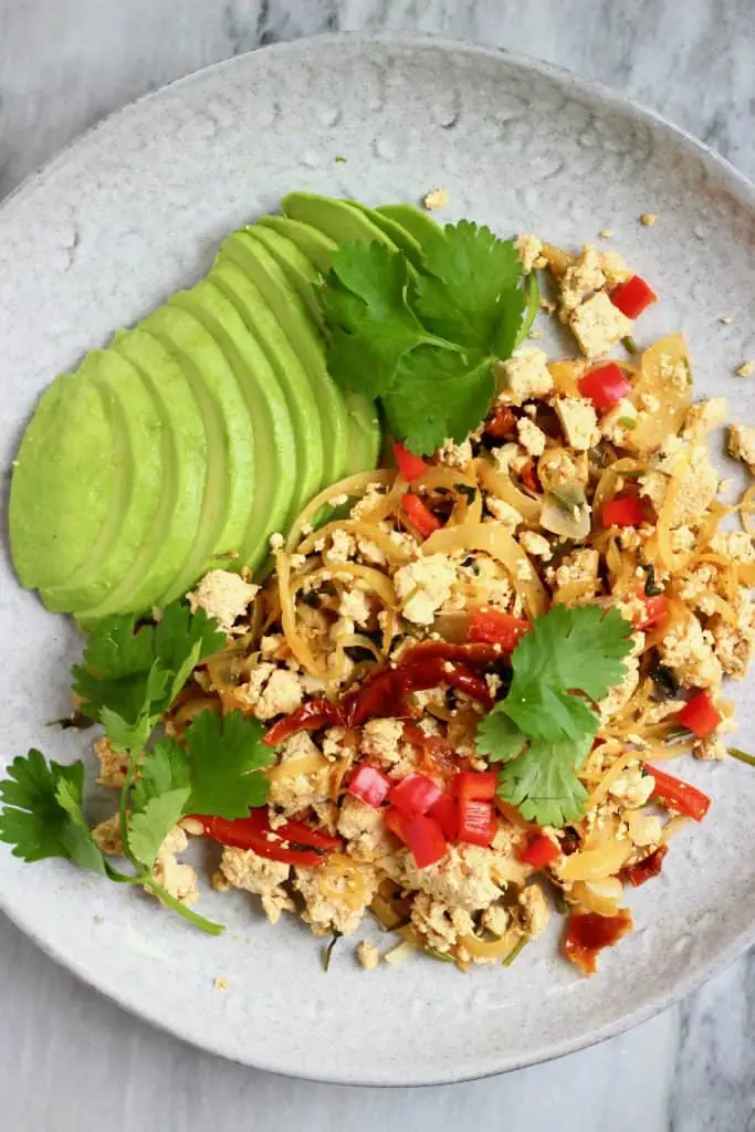 Scrambled tofu with red peppers and a sliced avocado on a grey plate against a marble background