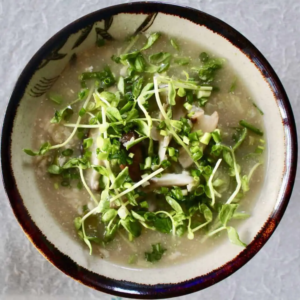 A bowl of brown rice miso soup topped with pea shoots in a white bowl with a dark brown rim against a white background