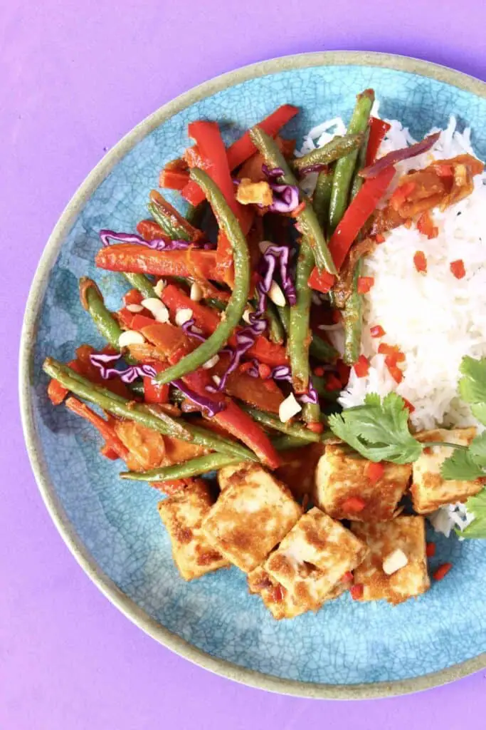 Brown cubes of tofu, sliced red pepper and green beans in a stir fry with a pile of white rice on a blue plate against a purple background