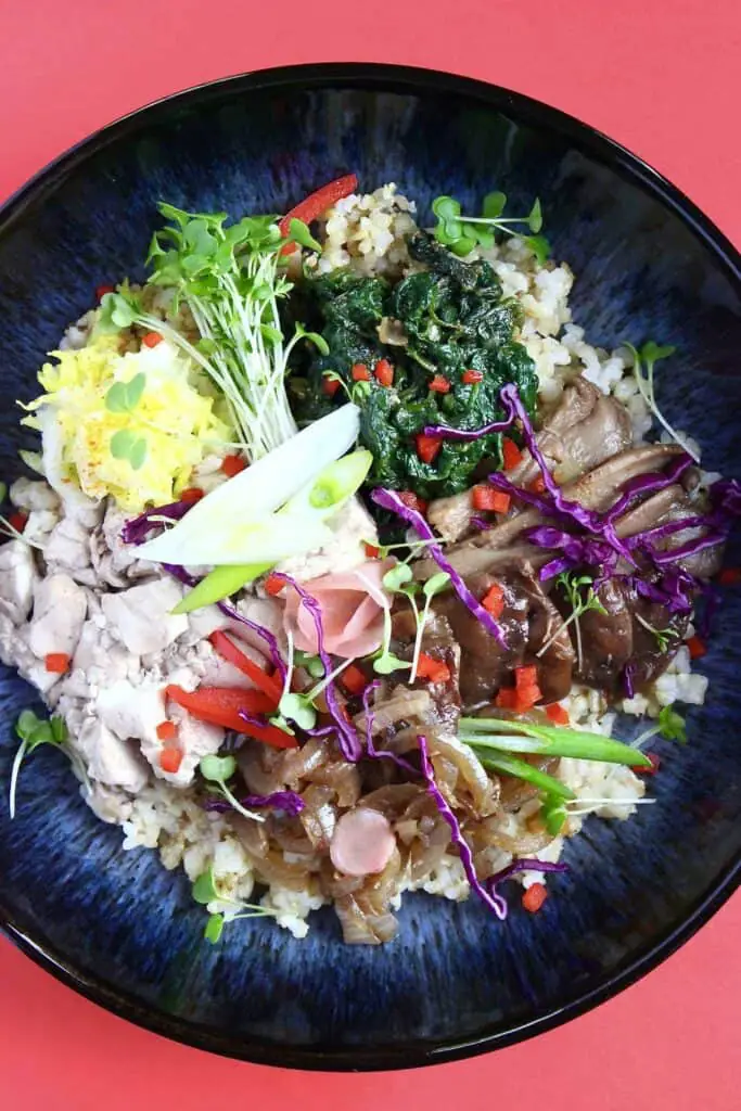 A bowl of rice topped with oyster mushrooms, and lots of different vegetables in a dark blue bowl against a red background