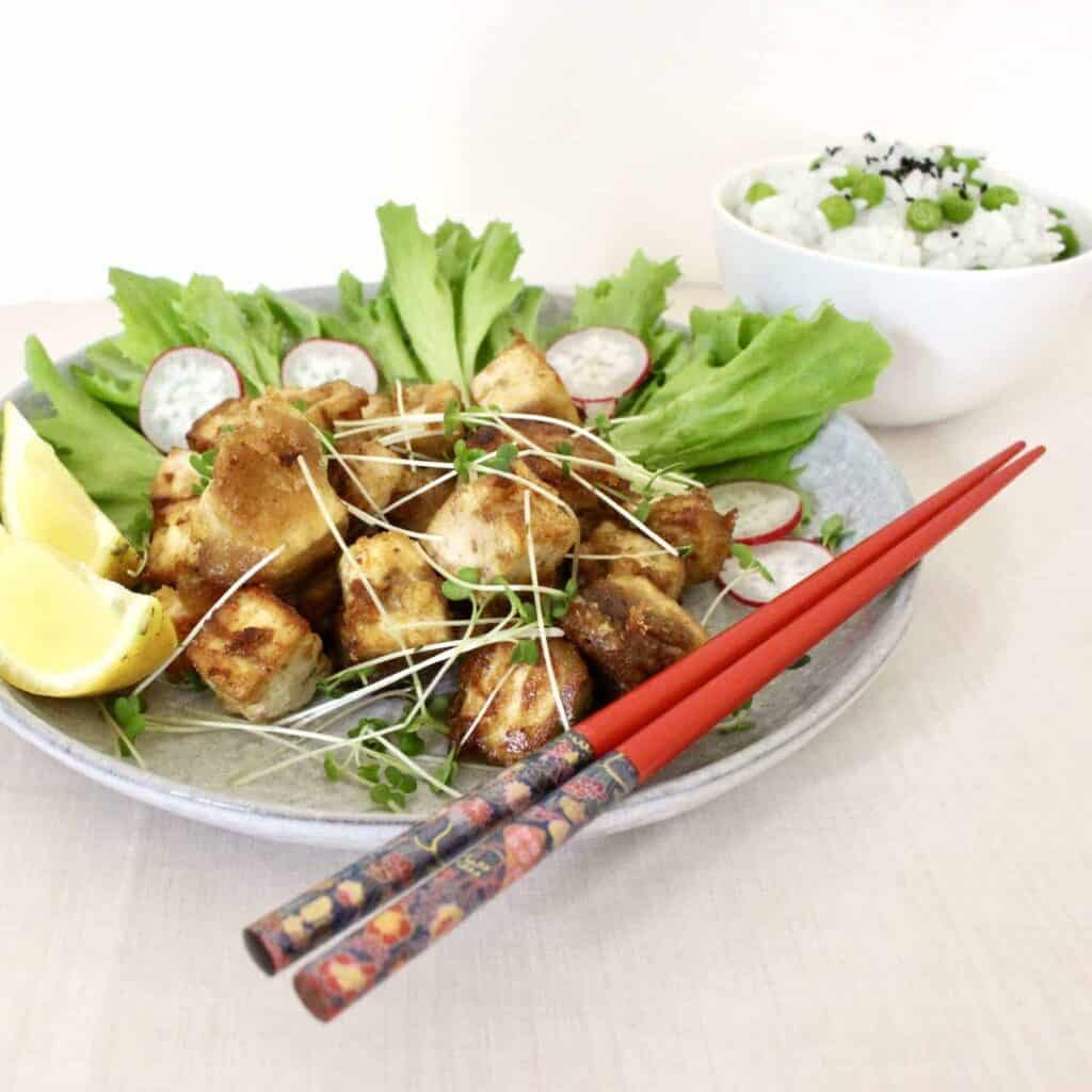 Pile of golden brown cubes of tofu with lettuce on a grey plate with red chopsticks