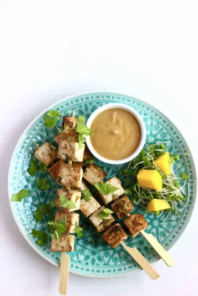 Tofu skewers on a green plate with a small white bowl of light brown sauce against a white background