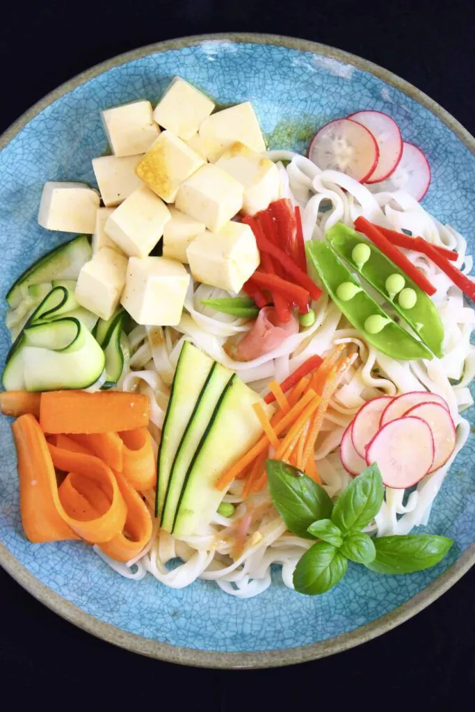Rice noodles, cubed tofu and raw vegetables on a blue plate against a black background