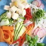 Rice noodles, cubed tofu and raw vegetables on a blue plate against a black background