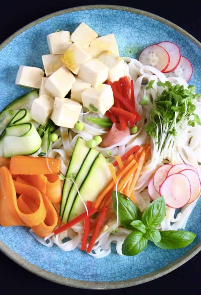 Cubes of tofu, carrot ribbons, zucchini ribbons, sliced red pepper, sliced radishes and fresh basil on a bed of rice noodles against a blue plate