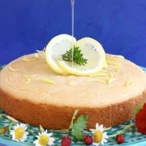 Gluten-free vegan lemon drizzle cake on a cake stand with a spoon drizzling syrup over it