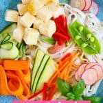 Rice noodles, cubed tofu and raw vegetables on a blue plate against a black background