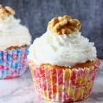 Two carrot cake cupcakes topped with creamy frosting and a walnut on a marble slab against a grey background