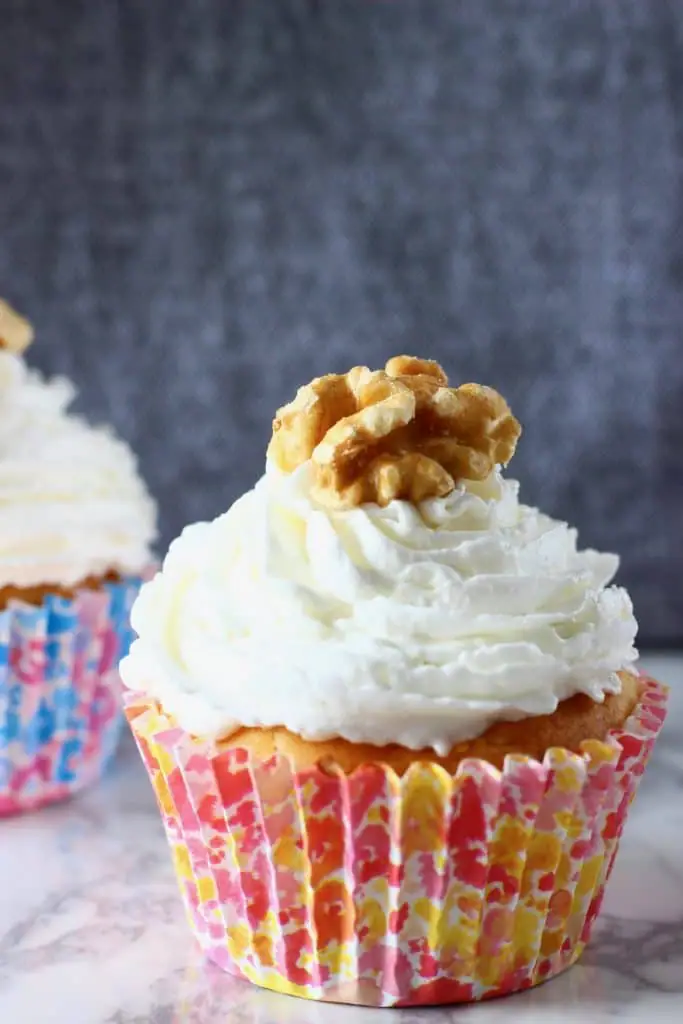 Two carrot cake cupcakes topped with creamy frosting and a walnut on a marble slab against a grey background