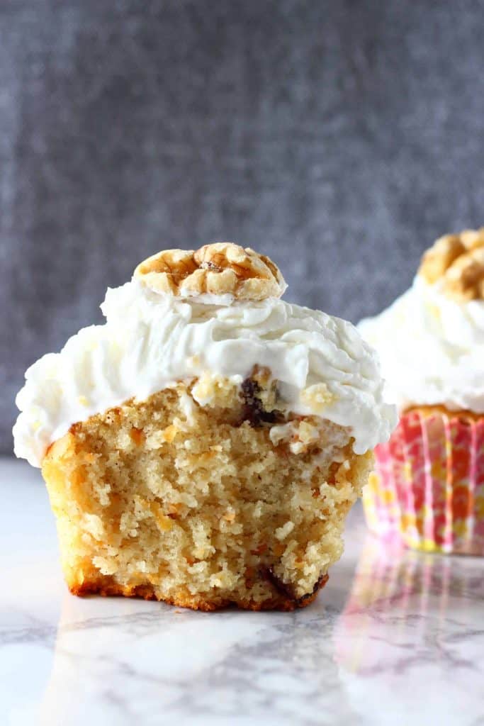 Two carrot cake cupcakes topped with creamy frosting and a walnut on a marble slab against a grey background