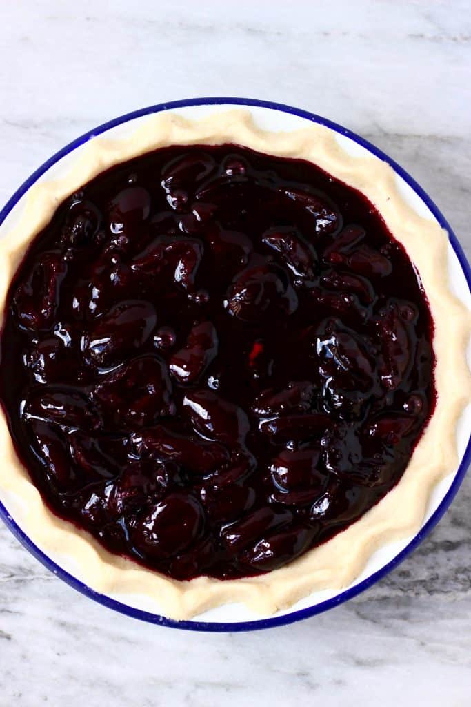 Cooked cherries in raw pie crust in a white pie dish with a blue rim against a marble background