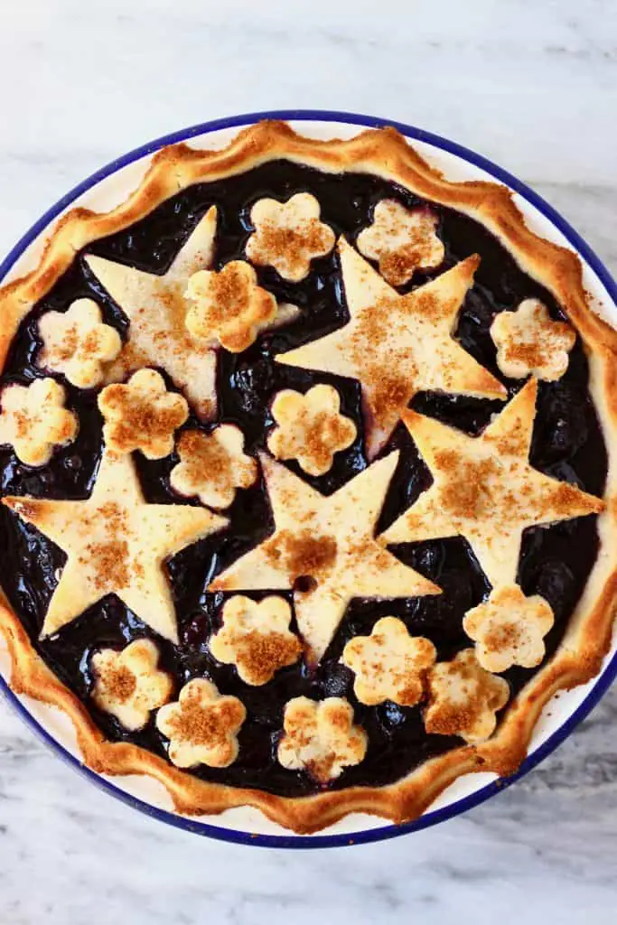 Cherry pie topped with stars and flowers in a white pie dish with a blue rim against a marble background