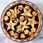 Cherry pie topped with stars and flowers in a white pie dish with a blue rim against a marble background