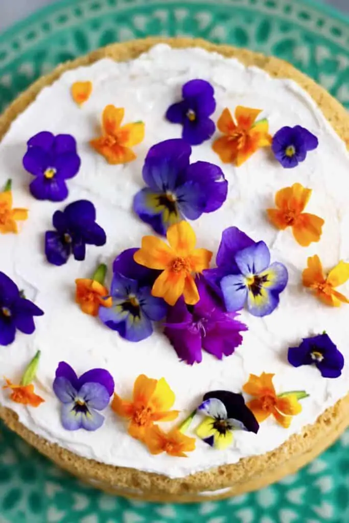 Photo of sponge cake topped with white cream frosting and orange and purple flowers on a green cake stand