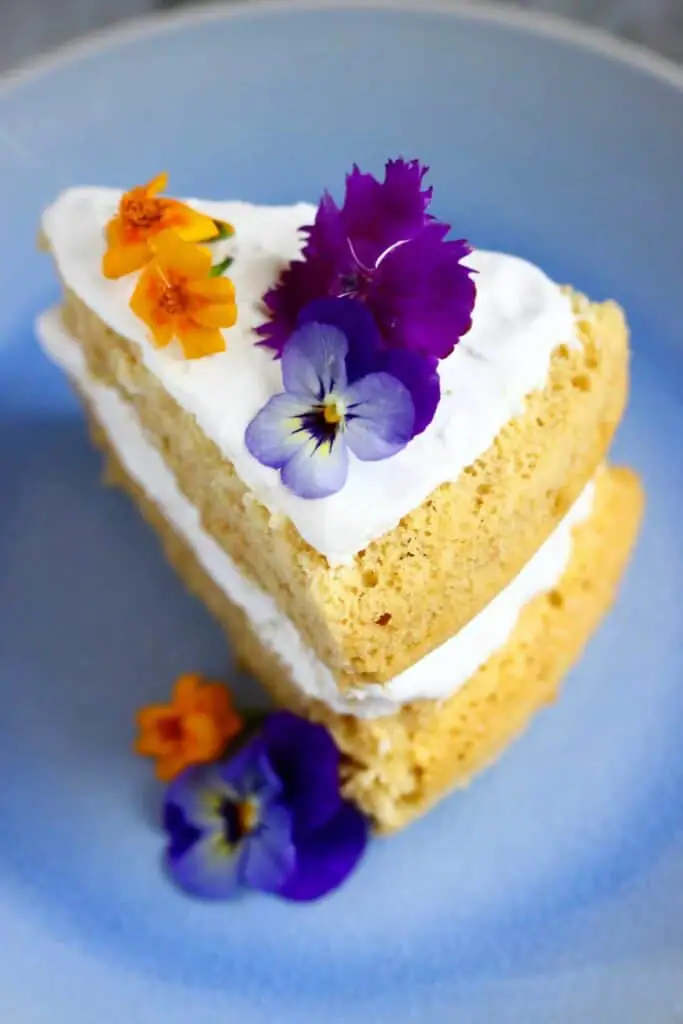 Photo of a slice of sponge cake sandwiched with white creamy frosting decorated with purple and orange flowers on a light blue plate