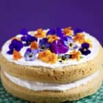 Photo of orange sponge cake sandwiched with creamy white frosting topped with orange and purple flowers on a green cake stand against a purple background