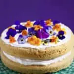 Photo of orange sponge cake sandwiched with creamy white frosting topped with orange and purple flowers on a green cake stand against a purple background