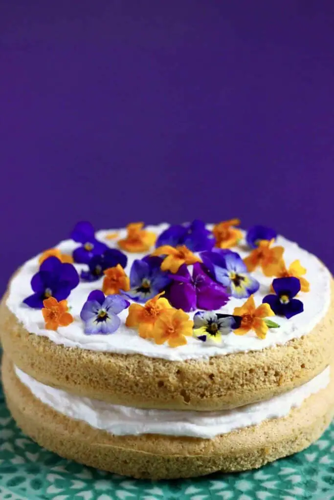 Photo of orange sponge cake sandwiched with creamy white frosting topped with orange and purple flowers on a green cake stand against a purple background