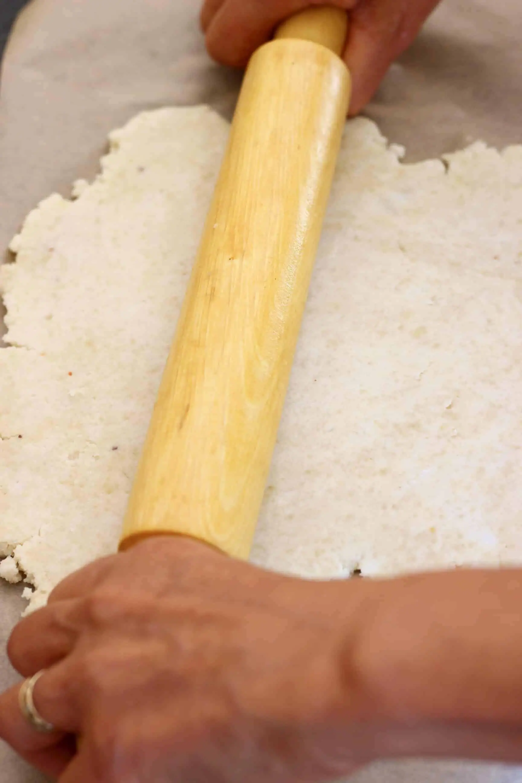 A sheet of brown baking paper topped with pastry dough with two hands and a rolling pin rolling it out