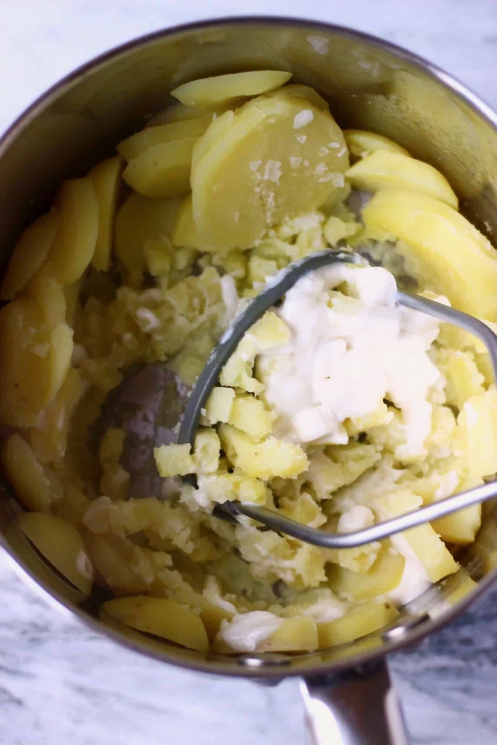 Slicked cooked potatoes and blended cashew cream in a pan being mashed with a potato masher