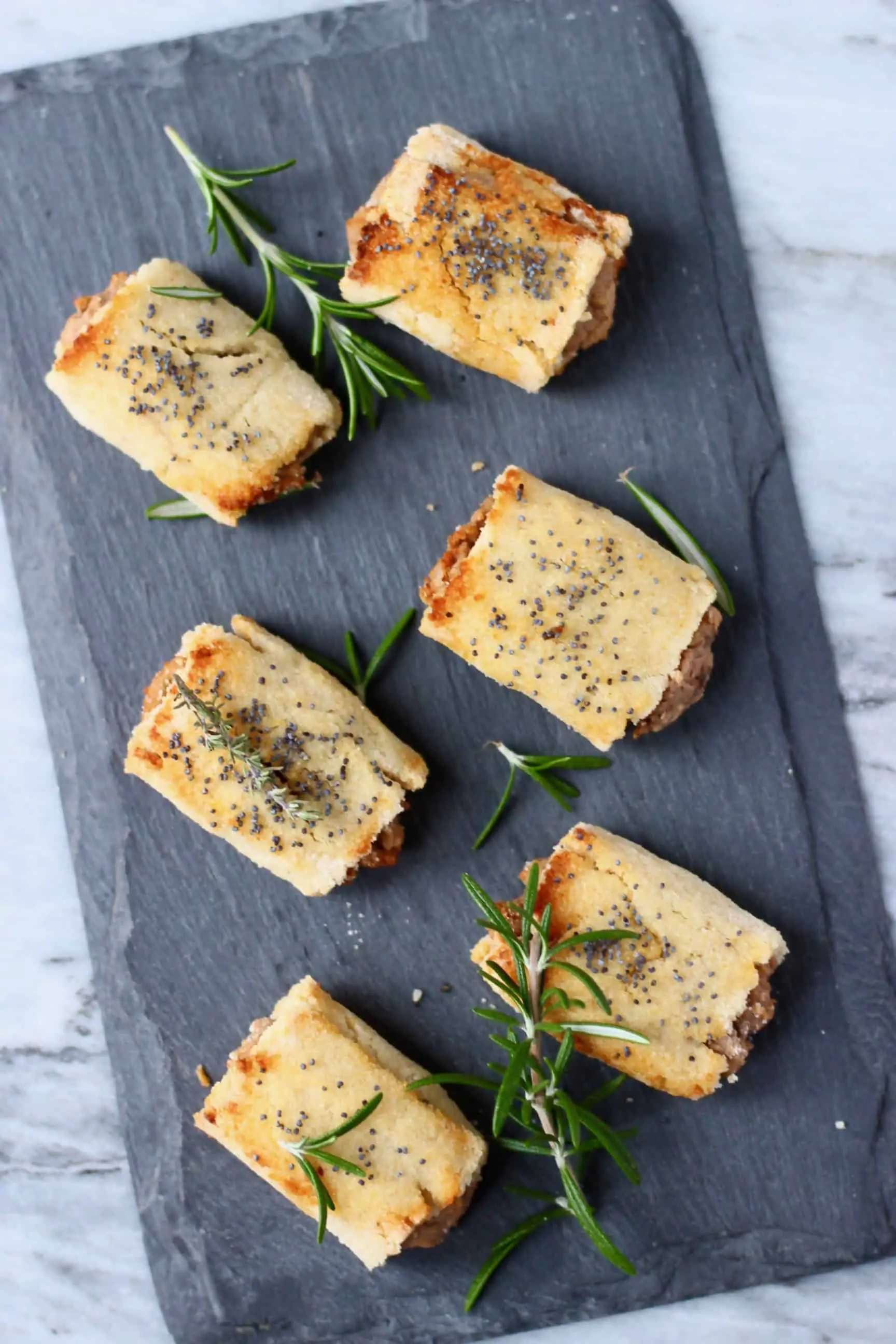 Six vegan sausage rolls on a black slab decorated with sprigs of rosemary