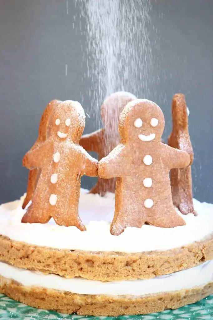 Photo of gingerbread sponge cake sandwiched with white cream topped with five gingerbread men cookies standing in a circle with a dusting of white icing sugar