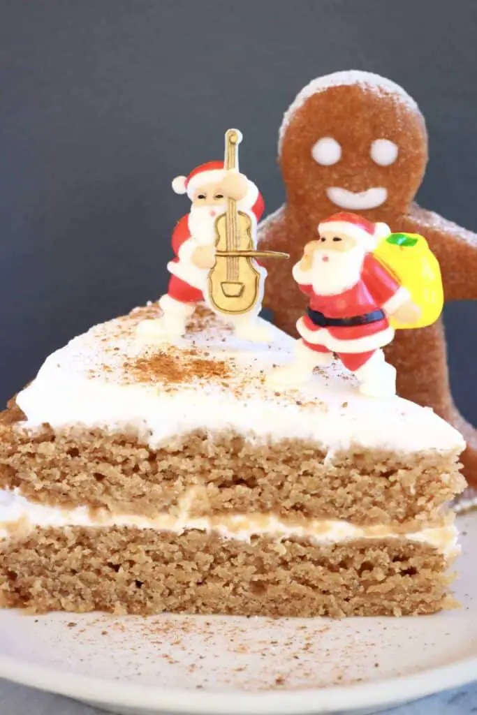 Photo of a slice of brown sponge cake sandwiched with white cream and decorated with plastic santa decorations with a gingerbread cookie behind against a grey background