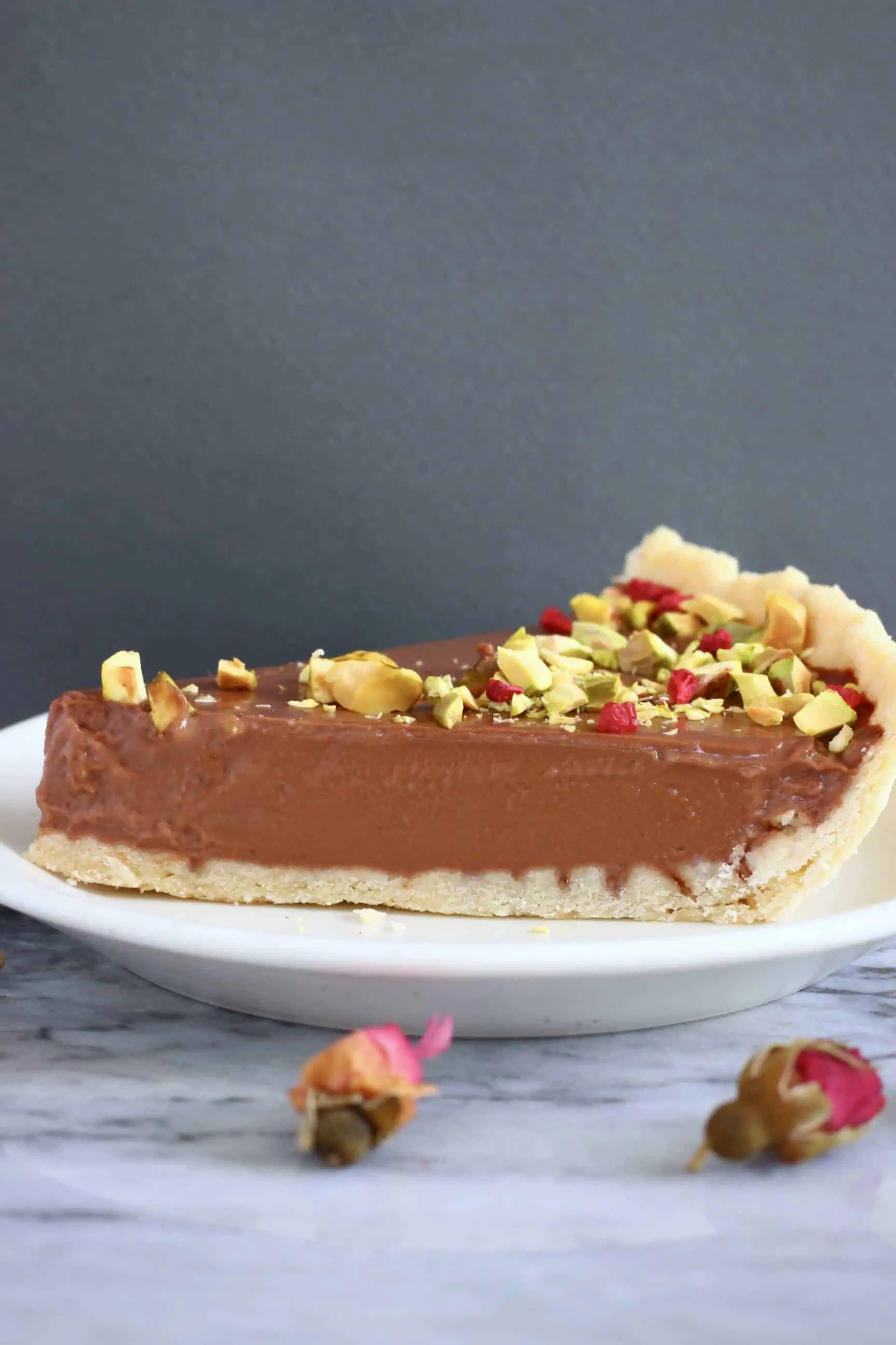 A slice of gluten-free vegan chocolate tart topped with chopped pistachios and freeze-dried raspberries on a plate