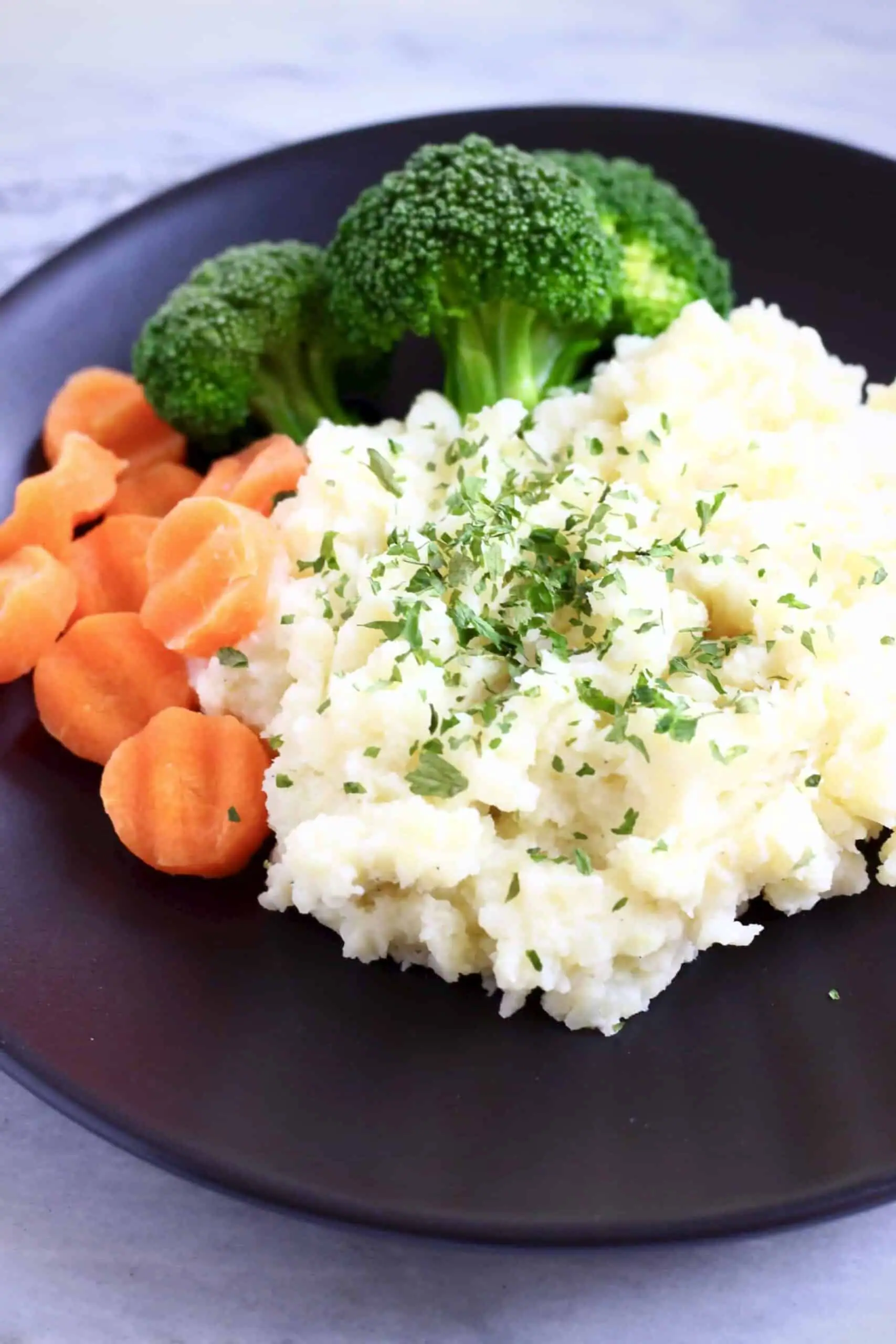 A pile of creamy vegan mashed potatoes sprinkled with green herbs with sliced carrots and broccoli on a plate