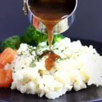 A pile of creamy vegan mashed potatoes topped with green herbs with sliced carrots and broccoli on a plate with vegan gravy being poured over