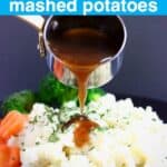 Photo of a pile of mashed potatoes topped with green herbs with sliced carrots and broccoli on a black plate with brown gravy being poured over in a silver saucepan against a dark grey background