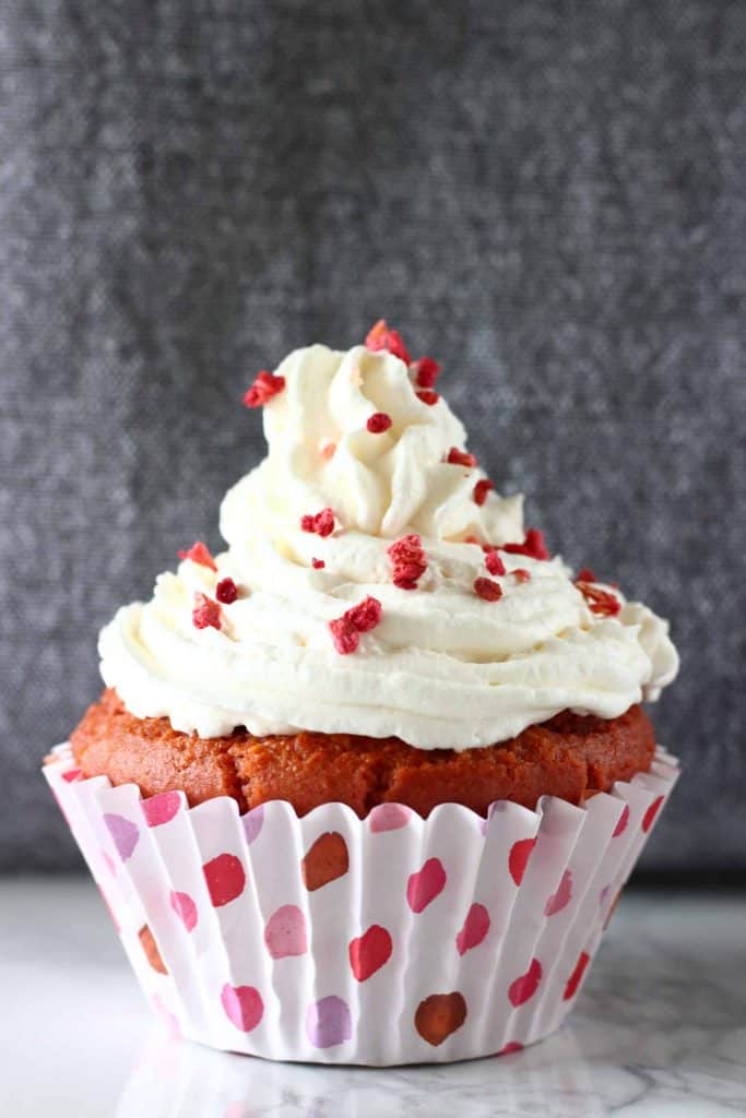 Red velvet cupcake topped with white frosting and freeze-dried raspberries on a marble slab against a grey background