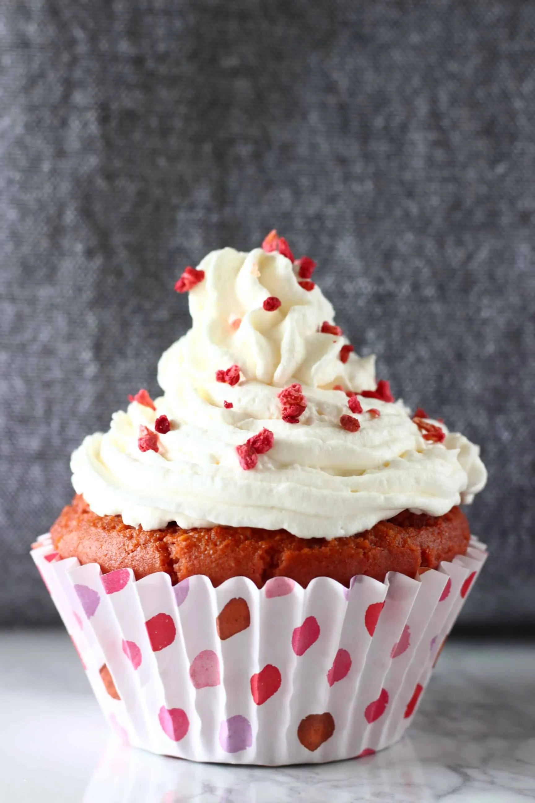 A gluten-free vegan red velvet cupcake topped with white cream cheese frosting and freeze-dried raspberries