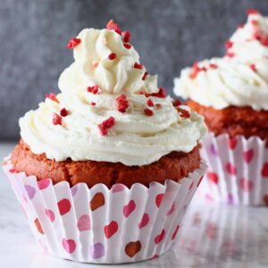 Two gluten-free vegan red velvet cupcakes topped with white frosting and freeze-dried raspberries on a marble slab against a grey background