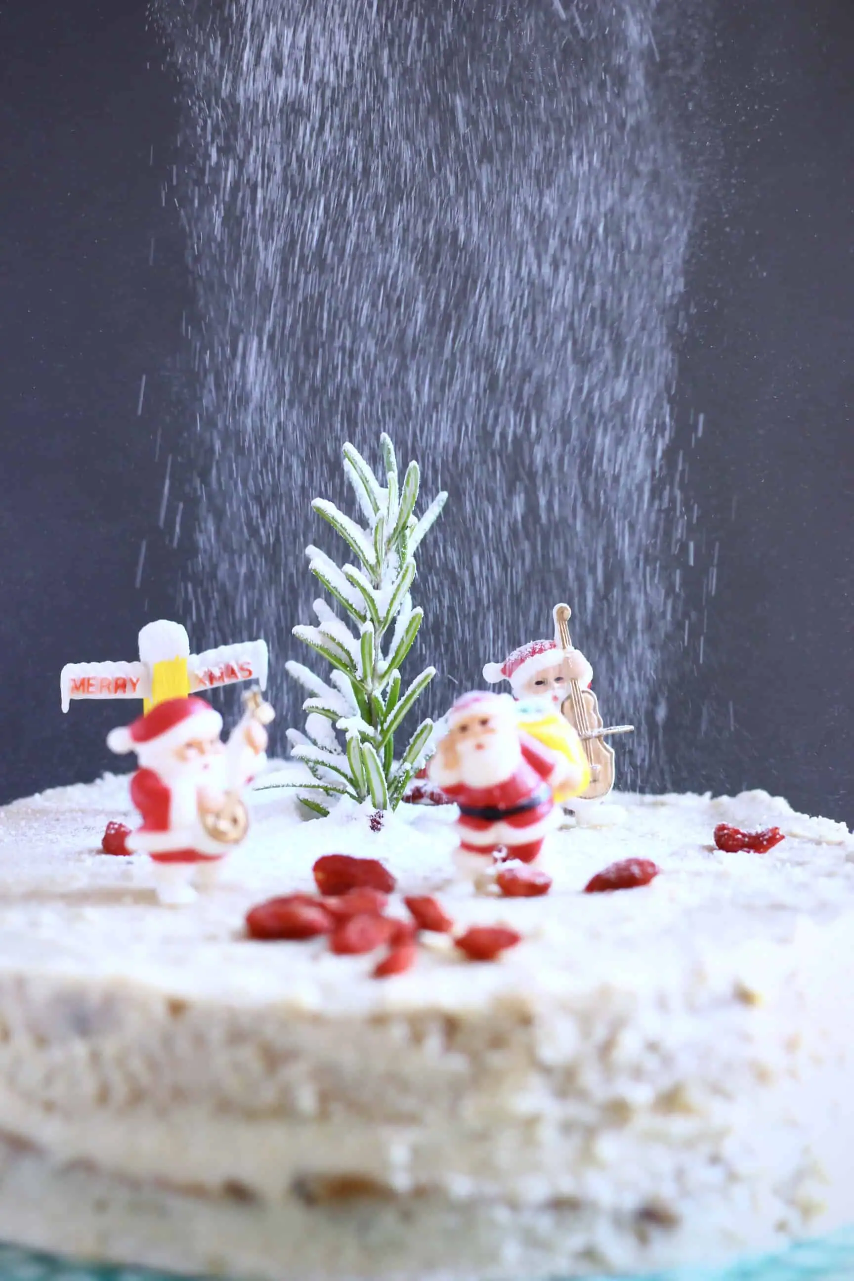 A gluten-free vegan Christmas fruit cake covered in white buttercream topped with plastic santas against a grey background with icing sugar being dusted over it