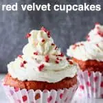Two red velvet cupcakes topped with white frosting and freeze-dried raspberries on a marble slab against a grey background