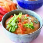 A blue bowl of orange vegan quest topped with green pepper and chopped coriander on a pink background with chopped vegetables and a dark blue bowl of tortilla chips