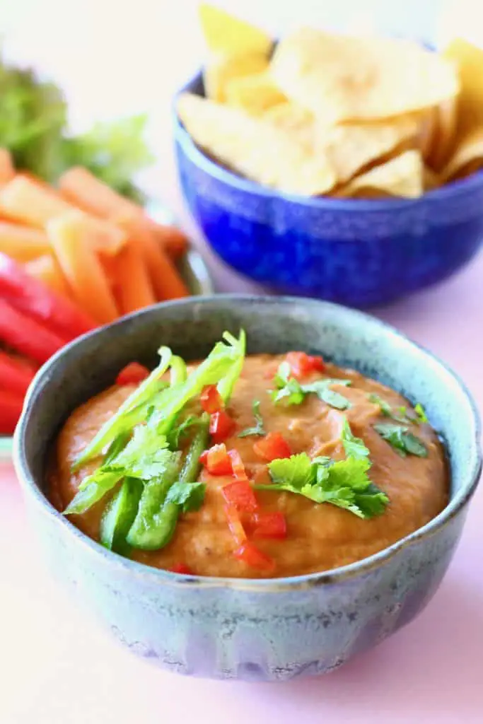 A blue bowl of orange vegan queso topped with green pepper and chopped coriander on a pink background with chopped vegetables and a dark blue bowl of tortilla chips
