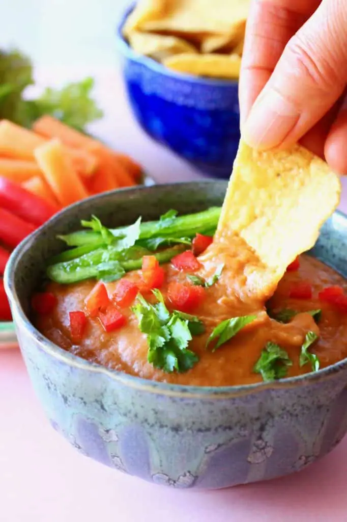 A blue bowl of orange vegan queso topped with green pepper and chopped coriander on a pink background with chopped vegetables and a dark blue bowl of tortilla chips with a tortilla chip being dipped into the queso